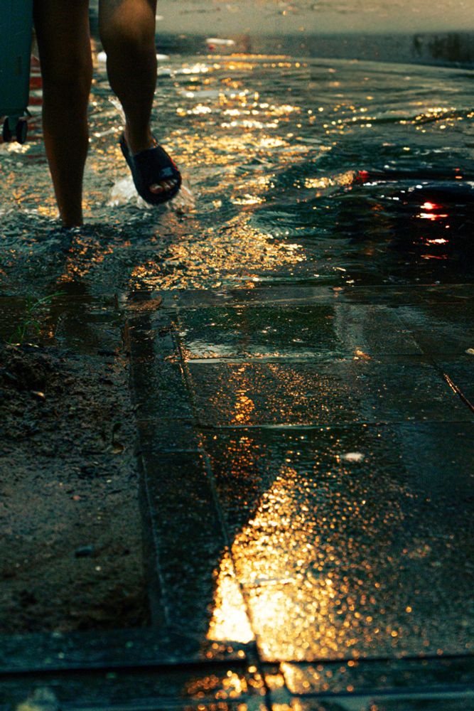 Close-up of legs walking through shallow floodwater on a city sidewalk, wearing blue sandals and pulling a suitcase. The wet pavement glows with golden reflections from streetlights.