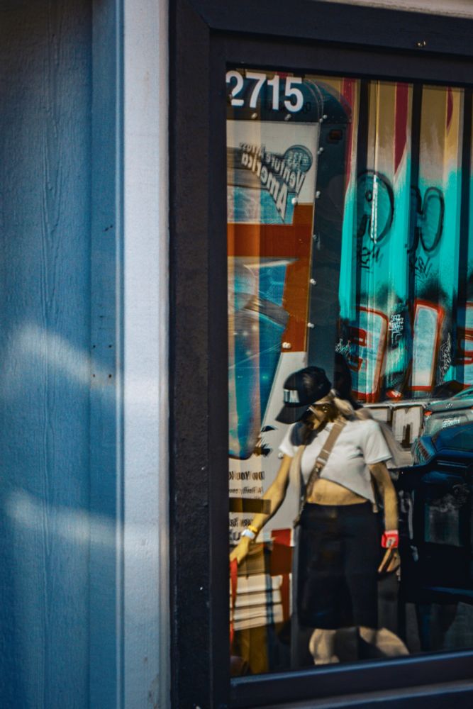 A woman in a cropped white shirt and black cap is reflected in a storefront window. Behind her reflection, bold graffiti and posters fill the background, creating layers of urban texture. Her face is partially obscured by reflection and angle.