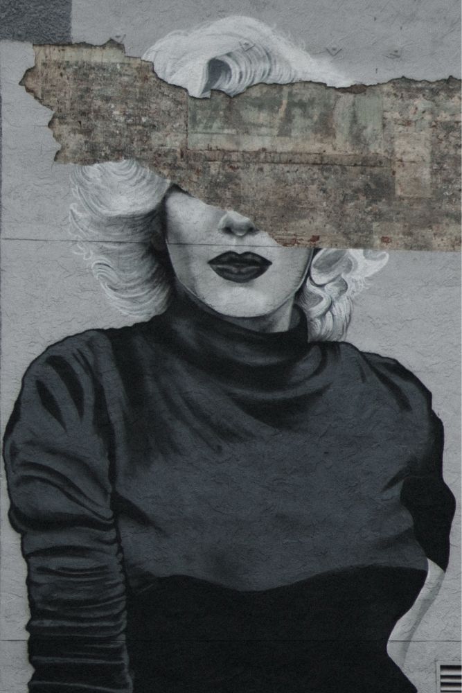 A black-and-white mural of Marilyn Monroe is painted on a textured wall. A large section of the building’s facade has peeled away due to storm damage, revealing rough, weathered material underneath. This missing piece covers Marilyn’s eyes, leaving only her lips and hair visible. The contrast between the smooth painted portrait and the jagged, deteriorated wall gives the image a fragmented and eroded look.