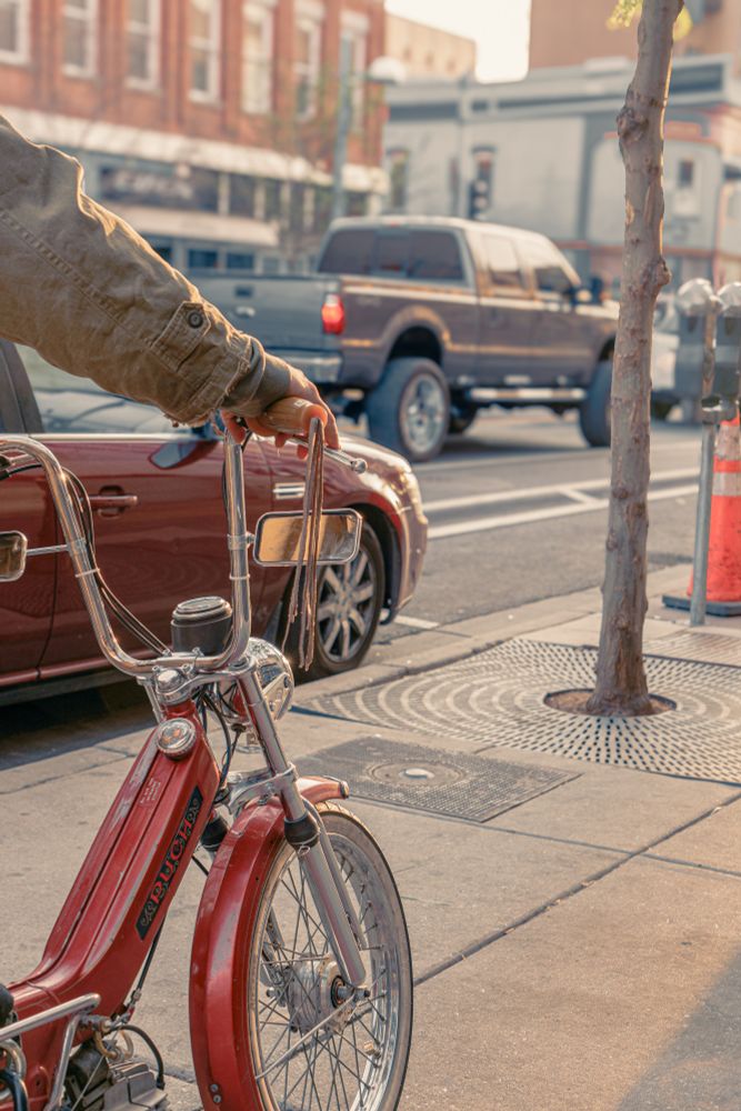 A close-up of a vintage, moped-style bicycle parked on a city sidewalk during golden hour. A person’s hand, wearing a rugged olive-green jacket, rests casually on the tall chrome handlebars. The sunlight catches the polished metal and red frame, casting warm highlights. In the background, cars pass through a lively urban street with red brick buildings and storefronts. A tree trunk and orange traffic cone sit unobtrusively on the right edge of the frame.
