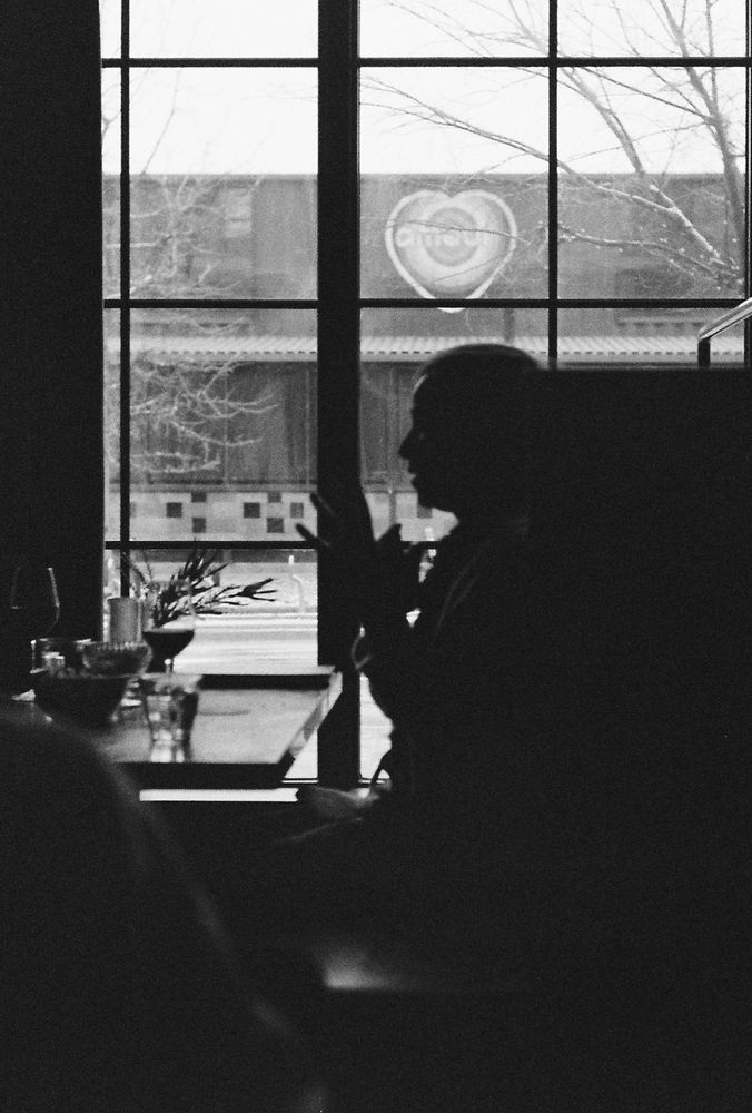 A black and white photograph of a silhouetted person sitting at a café table, mid-gesture, framed against a large window. Outside, a heart-shaped sign with the word “Choose” is visible on a building across the street. The table holds glasses and a sprig of greenery, suggesting an intimate, contemplative moment.