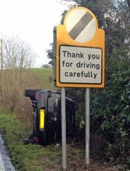 A road sign saying "thank you for driving carefully" and beyond that a crashed car turned on its side.