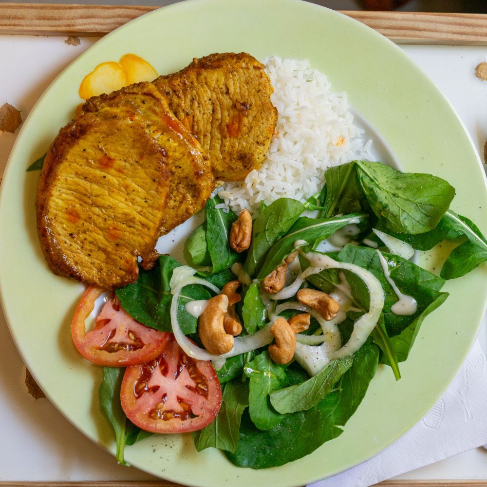 Prato de comida com lombo, arroz, salada de rúcula, cebola, tomate e castanhas de caju 