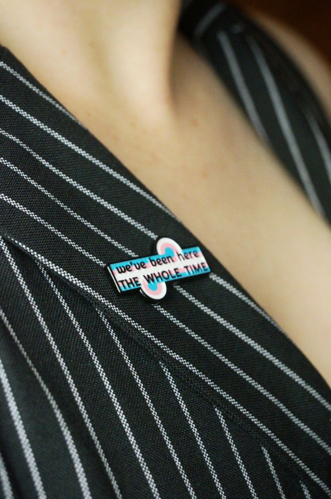 The trans game changer pin on a pinstripe lapel. The pins says "we've been here the whole time"