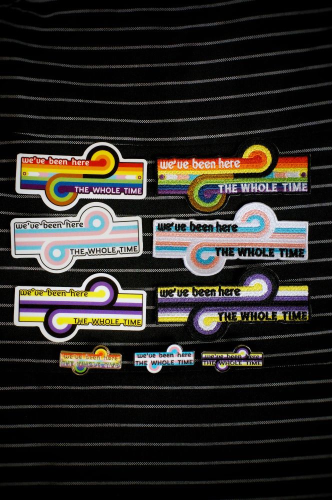 Stickers, embroidered patches, and enamel pins in LGBTQ+, trans, and non-binary flags colours. The designs are the Game Changer loop-de-loop pattern and the words "we've been here the whole time"