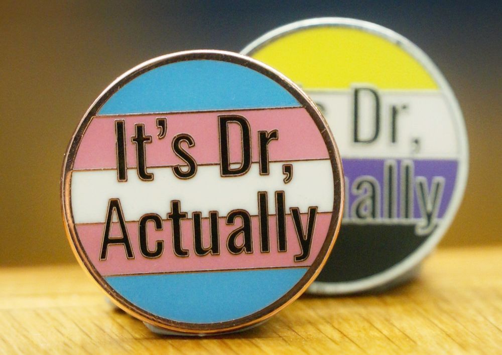 Two circular enamel pins with the text "It's Dr, actually" in black. The rest of the design is the transgender pride flag on one pin and the non-binary pride flag on the other.