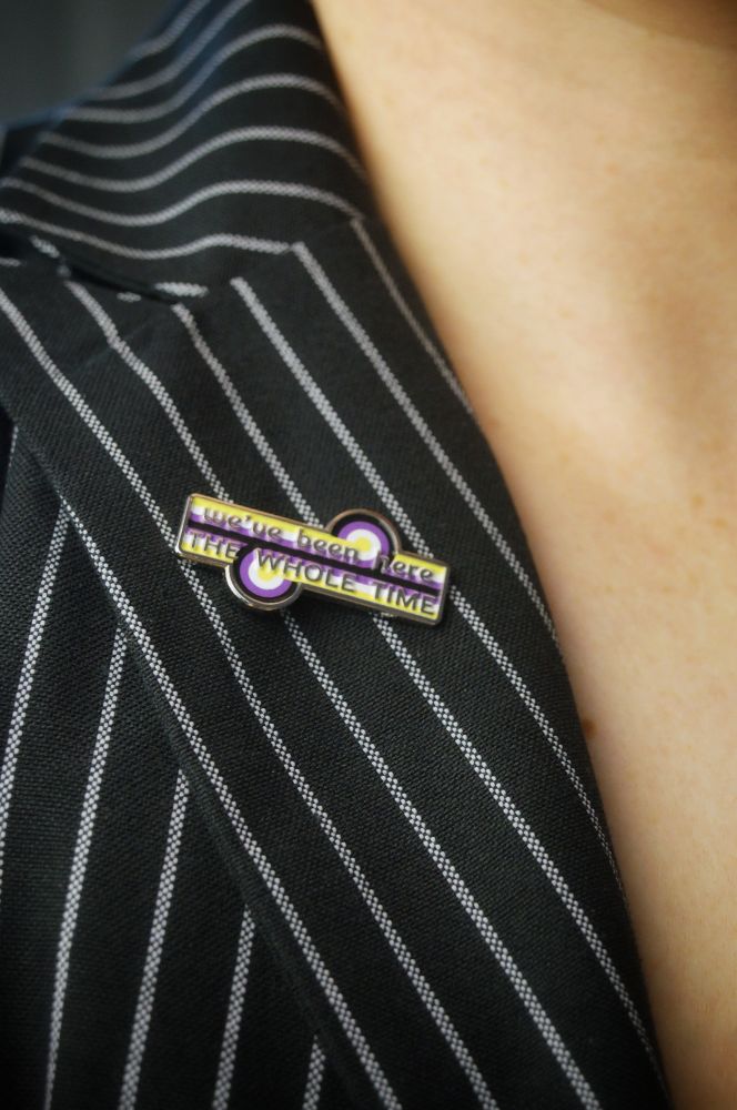 The non-binary game changer pin on a pinstripe lapel. The pins says "we've been here the whole time"