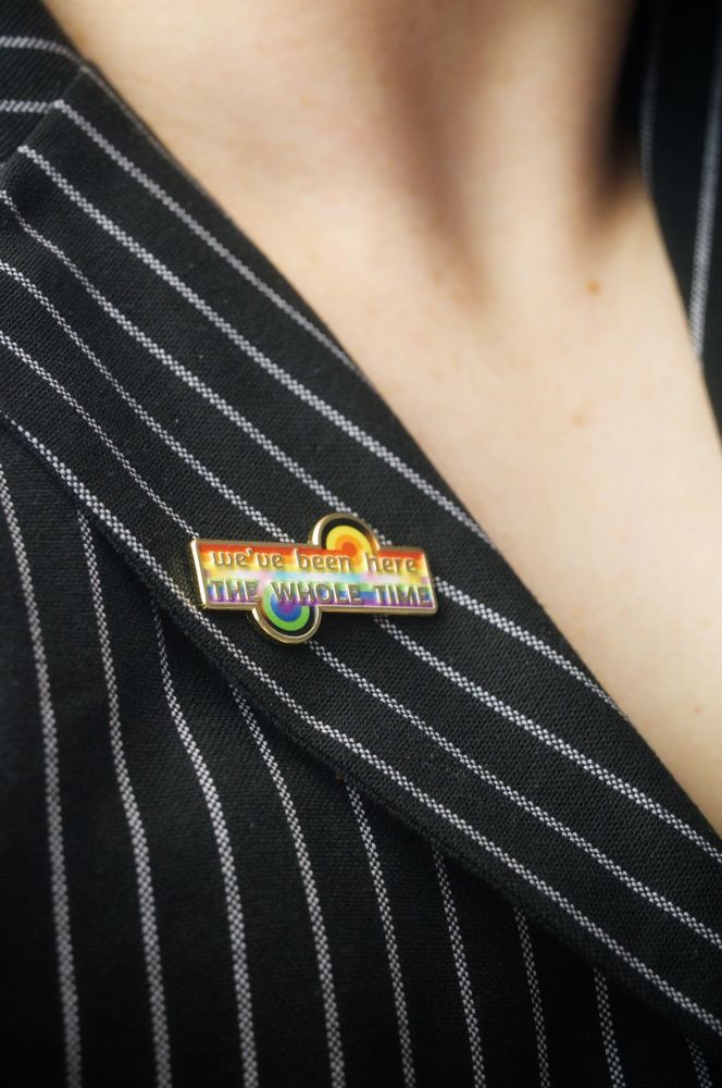 The LGBTQ game changer pin on a pinstripe lapel. The pins says "we've been here the whole time"