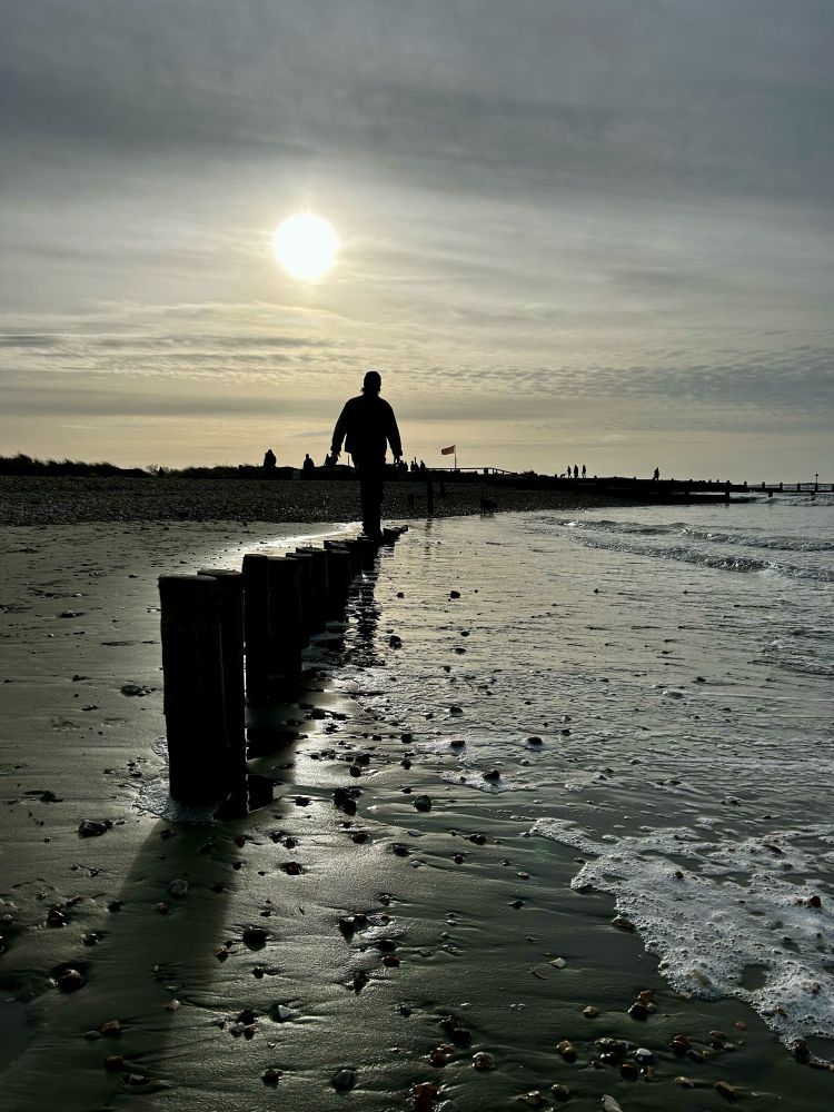 A young man silhouetted against a low winter sun walks atop some old groyne posts jutting from the beach at West Wittering, Sussex. Boxing Day 2023.
