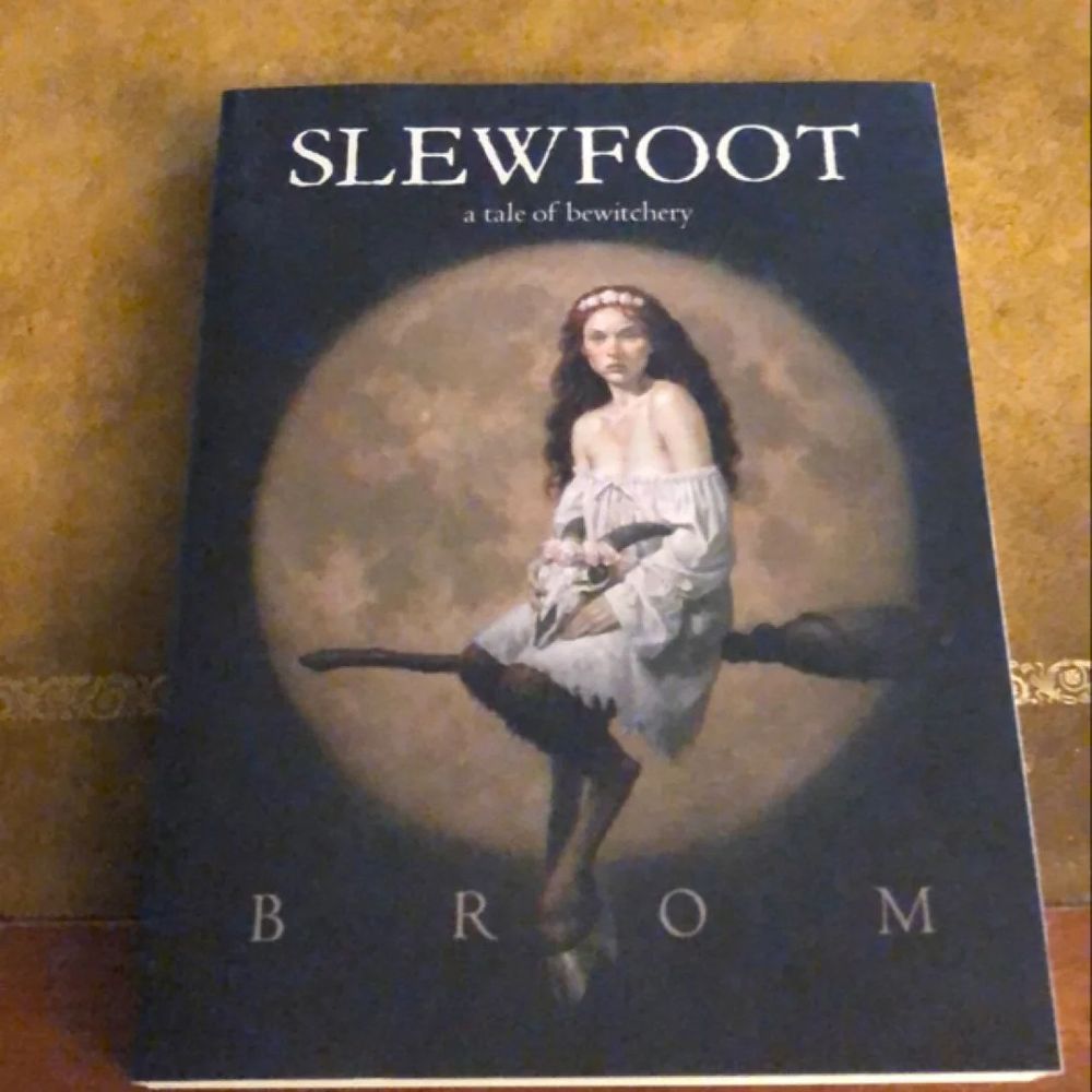 An image of a book-- SLEWFOOT a tale of bewitchery by Brom. 
The cover image is a cloven hooved witch riding a broom. She wears a chemise & a crown of flowers. She is young, beautiful w/ red hair. She holds a flower crowned goat skull in her lap.