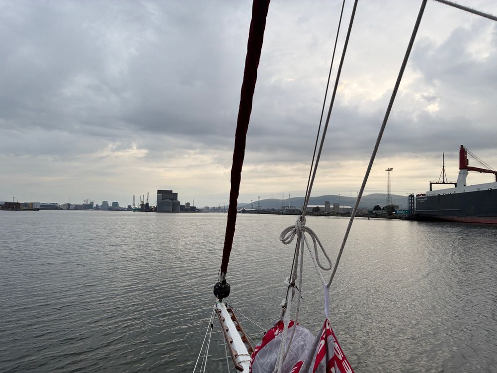 Entering the port of Belfast. It’s clearly and industrial working port. Several container ships can be seen berthed, including on quite close to the right. Indigo’s bowsprit in the foreground. It’s just before dusk and overcast.