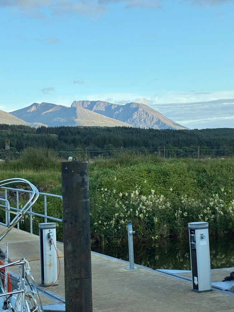 Ben Mevis, seen from Indigo’s pontoon on the Caledonian Canal. Some of the pontoon to be seen in foreground. The peak lit by late afternoon sun in the distance. Blue skies, and a forest between.