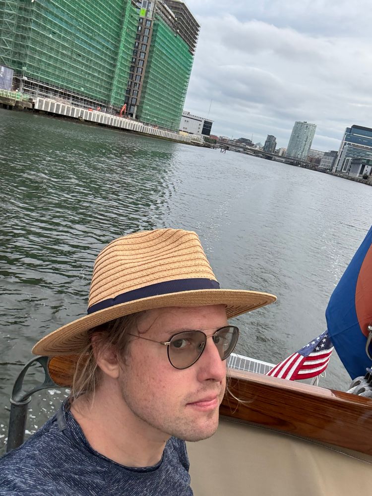 Your  correspondent in a side eye selfie from the stern of Indigo. Two green-glass modern building in background, on the right bank of the Lagan River (which forms the base of the harbour, and on which sits the marina). Center background, the several tall buildings of Belfast’s CBD. Indigo and I are leaving Belfast.