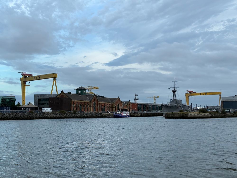 Looking back at the shipyard of Harland & Wolff while motoring through Belfast Harbour. The marina is just out of frame to the right. The two big yellow cranes - nicknamed Samson and Goliath - are a landmark of the Belfast Harbour skyline. Harland & Wolff built the titanic in the now filled slipway near the right edge of the photo! In the center, framed between the two gargantuan cranes, a WWI destroyer, the HMS Carloline, permanently moored as a museum ship. Skies are grey and partly cloudy. Water is calm.