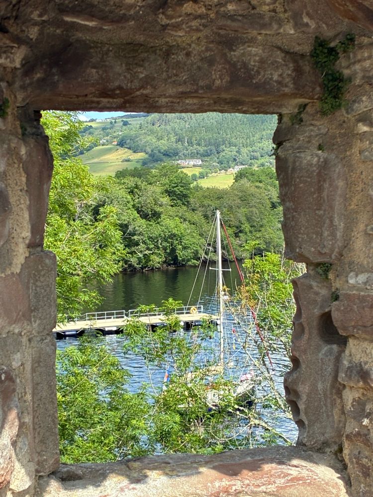 Indigo seen anchored close to shore and a pier. The shot is framed by a ruined castle window, and Indy is partially obscured by tree branches. It’s a sunny day and the waters, and distant green fields, shine.