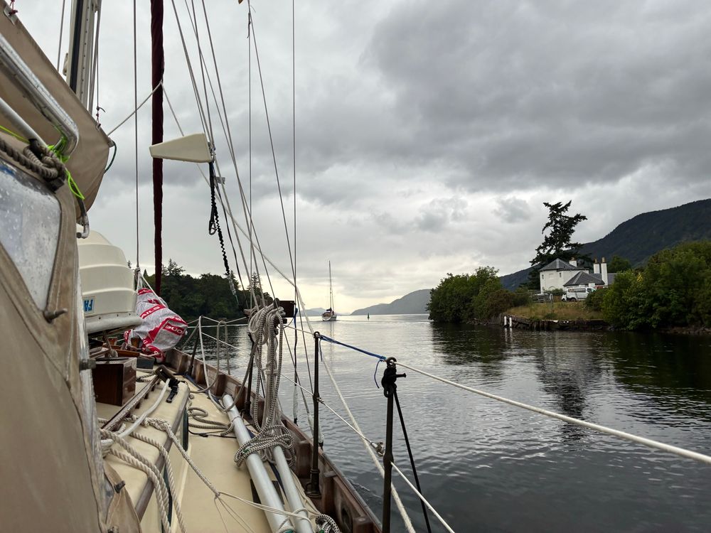 Entering Loch Ness out of the first part of the canal/River Ness. A narrow lake in front, a house to the right, two buoys and another sailboat ahead. The sky threatens rain but is suited to the landscape.