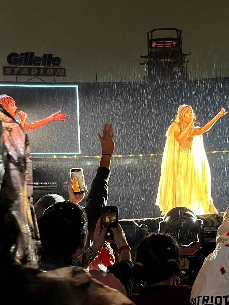 A photo of Taylor Swift brightly lit wearing a white flowy dress on stage during the folklore set of the eras tour in the pouring rain. Behind her is a big screen with Gillette stadium logo above it and in froths foreground of the photo are many fans with cameras out