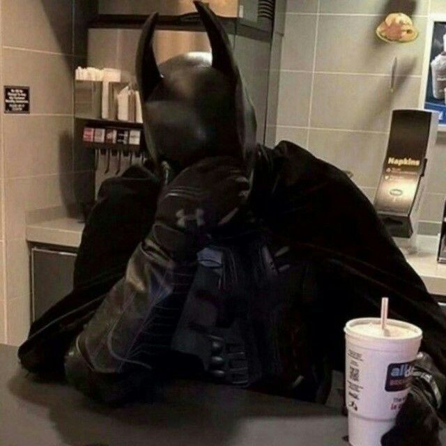 Batman cosplayer with one hand covering eyes, as if disappointed or brainfreeze, the other holding a fast food drink. Soda fountain in background.