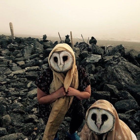 2 people wearing creepy barn owl masks, with a scarf draped over their heads, against a dreary rocky background, that really makes them look like survivors in rubble...
