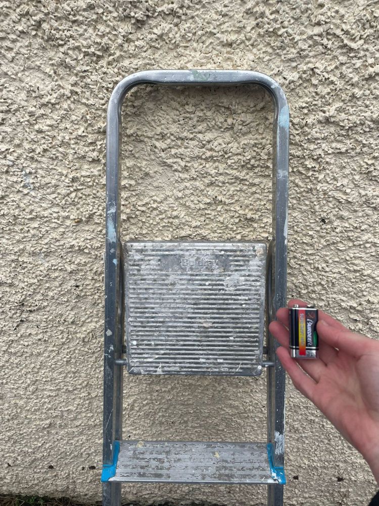 a photo of a step ladder leaning against a wall and a hand holding a 9v battery. 