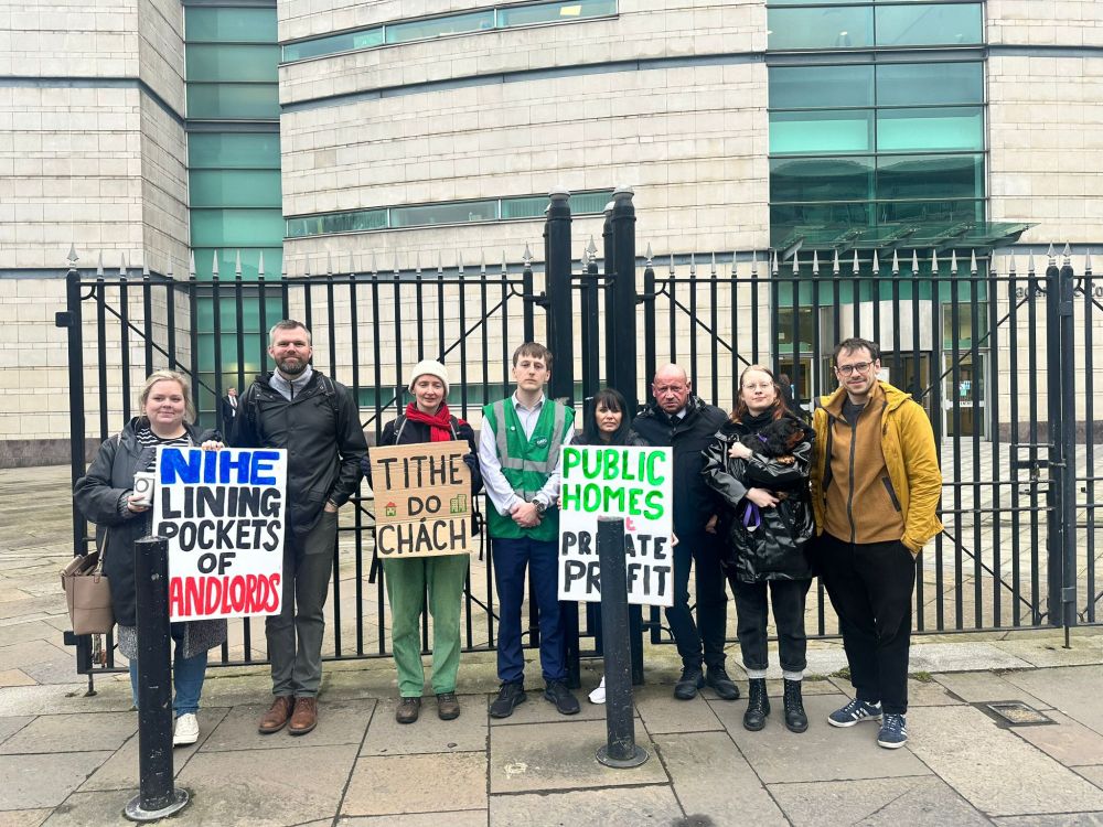 a photo of people standing in front of iron railing outside a court building. they are holding placards reading (left to right): NIHE lining pockets of landlords, Tithe do Chách, Public Homes not Private Profit