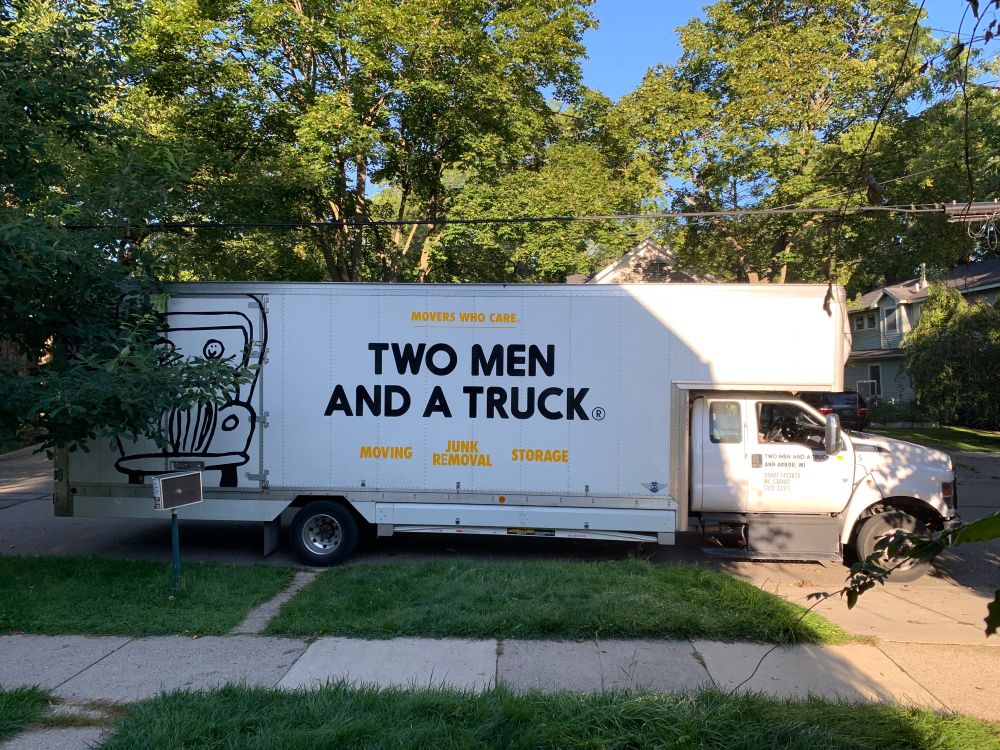 The background of the photograph is a sunny morning on a tree-lined street. Centered in the image is a white moving truck with the company’s name “Two Men and a Truck” printed on the side.