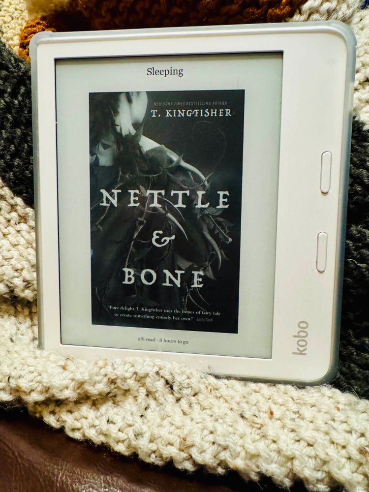 A Kobo on a blanket, currently in sleep mode. It’s showing the cover of “Nettle & Bone” by T. Kingfisher