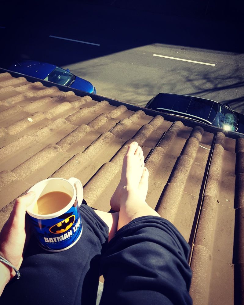 Foto meiner Beine, die übereinandergeschlagen sind und auf den Dachziegeln eines Dachs liegen. In der linken Hand eine Kaffeetasse mit Batman-Aufdruck. Bei sonnigem Wetter, weiter unten ist Straße zu sehen.