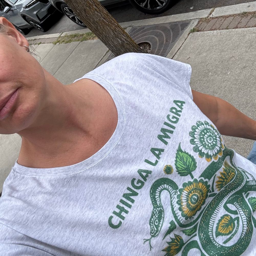 Woman wearing a white tshirt with an illustration of a green snake among green and yellow flowers with the words “chinga la migra” fuck ICE!