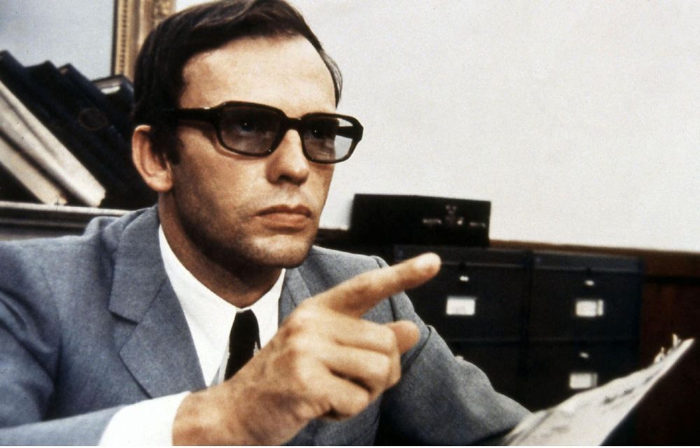 screen : Jean Louis Trintignant assis à un bureau, un papier à la main, il pointe son doigt vers quelqu'un
