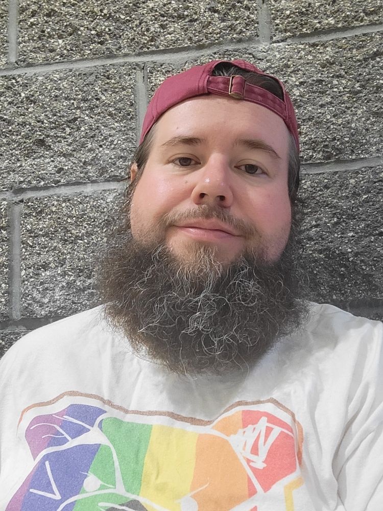 Man in a baseball cap against a cinder block wall. The T shirt is white and features an artist's creation of his logo with the modern pride inclusive flag.