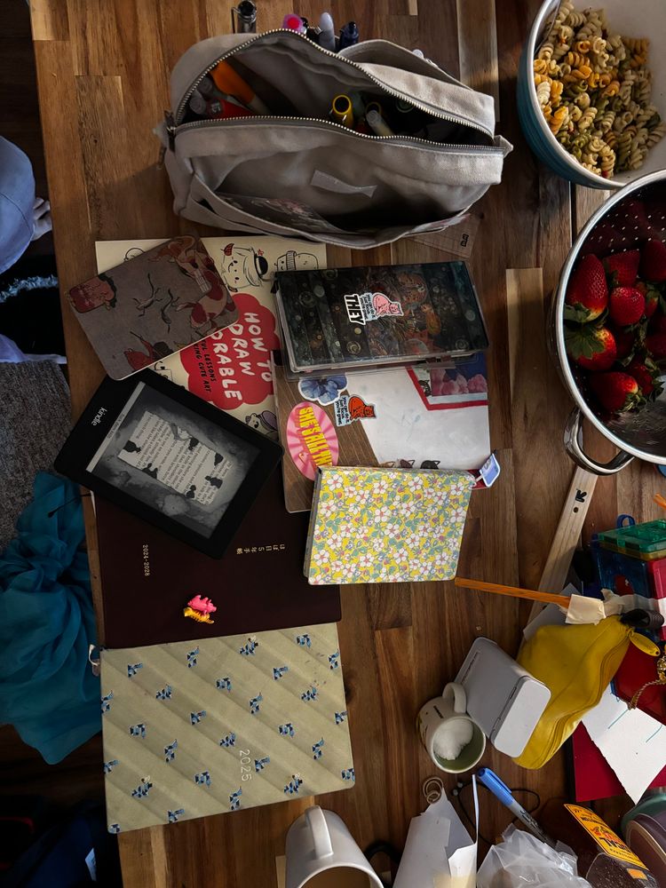 Photo of several diaries spread out on a kitchen table with a kindle and bag holding various fountain pens. There is a bowl of red strawberries near the top and a lot of clutter surrounding. 
