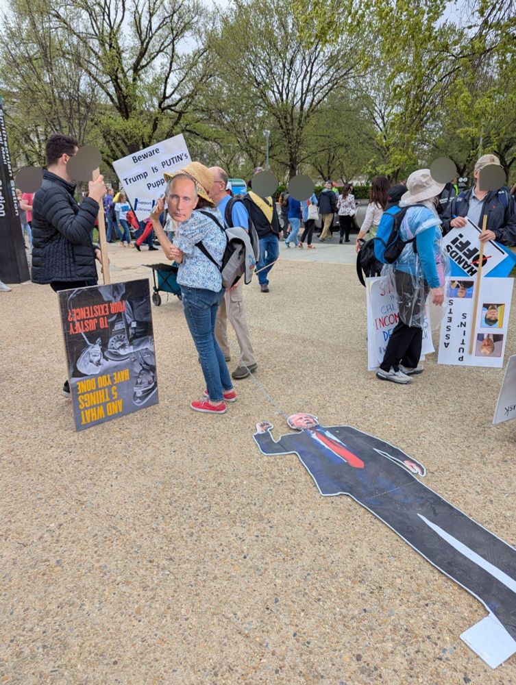 Protester wearing holding a Putin mask in front of their face dragging a cardboard Trump by a chain around his neck. Other protesters stand in the background