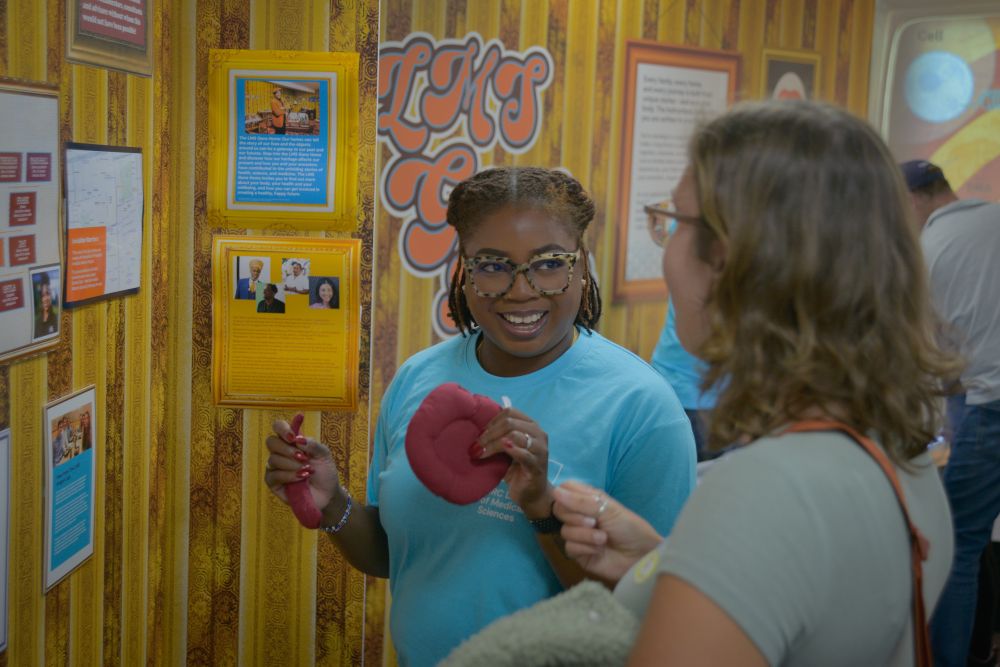A woman holding a model of 2 red blood cells, one affected by sickle cell and one unaffected, smiles while talking to another woman
