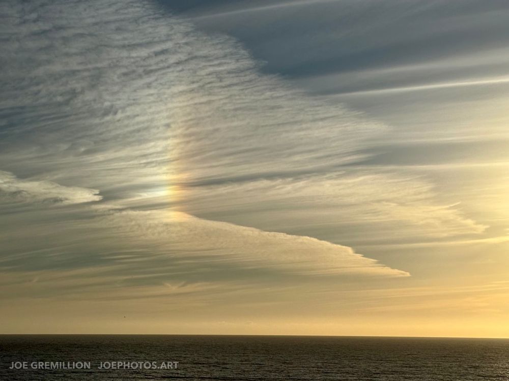 Scene of the Pacific Ocean at sunset. The sky dominates the view, filled with wispy clouds. A sun halo creates a spectral glow amidst the clouds. The foreground consists of the dark, tranquil water of the ocean. There are no people or any specific objects, only the vast expanse of nature.