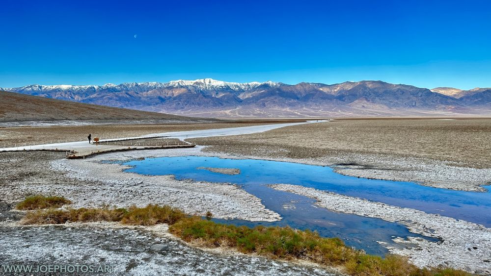 A lone figure stands on a wooden walkway above a reflective pool of water. The stark, white salt flats stretch out before them. In the distance, snow-capped mountains loom with peaks sharp against the horizon.