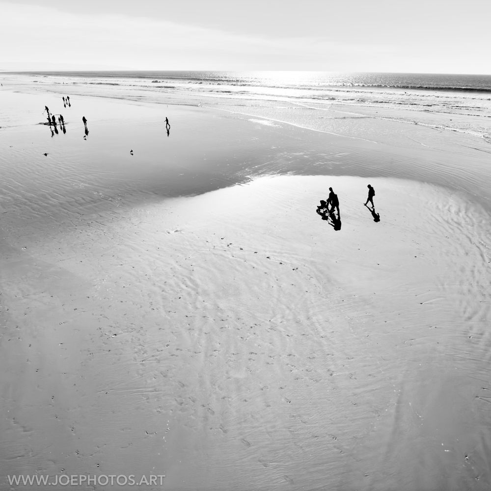 The scene unfolds on a vast, monochrome beach, with the ocean stretching out to the horizon under a bright sky. Silhouetted figures dot the landscape, including a family strolling with a stroller near the foreground and other individuals scattered along the shoreline. The smooth, wet sand reflects the sky and the figures, creating a mirror-like effect.