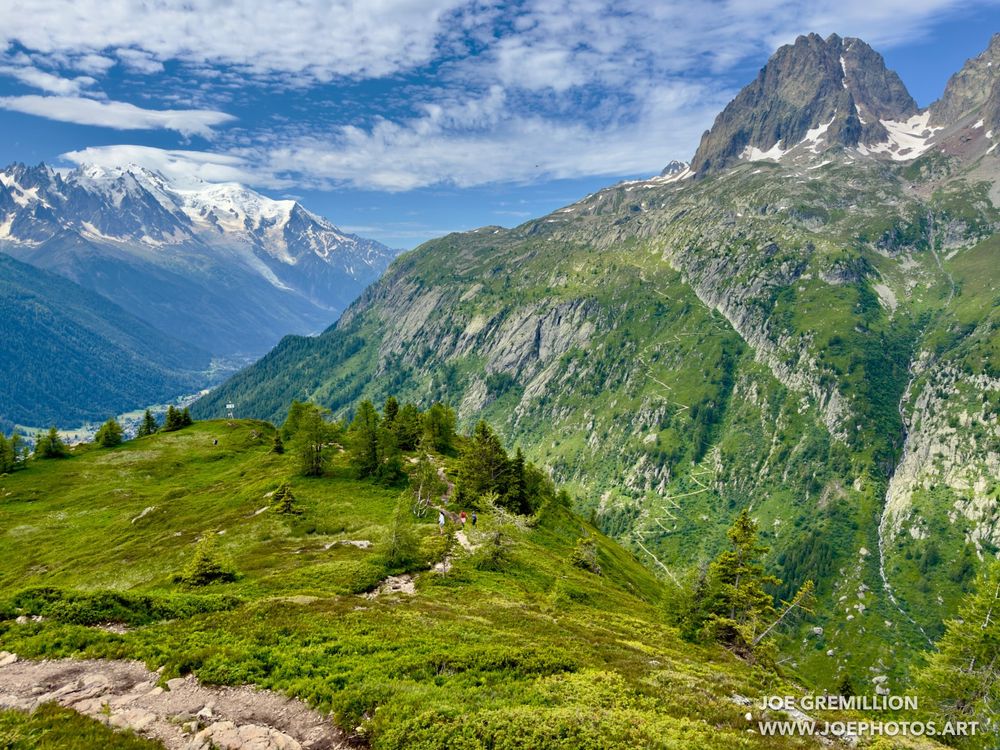 The landscape north of Chamonix. The foreground is lush with green vegetation and rocky paths, while the majestic snow-capped mountains loom in the background. The vast, open sky is dotted with clouds, enhancing the natural beauty of the French Alps.