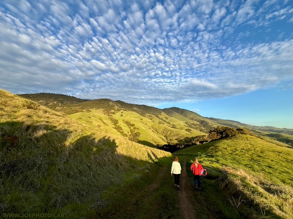 Two hikers walking along a dirt path with rolling green hills in the background. The near sky is filled with a smattering of clouds and clear in the distance. Sunset light is cast upon the hills and path.