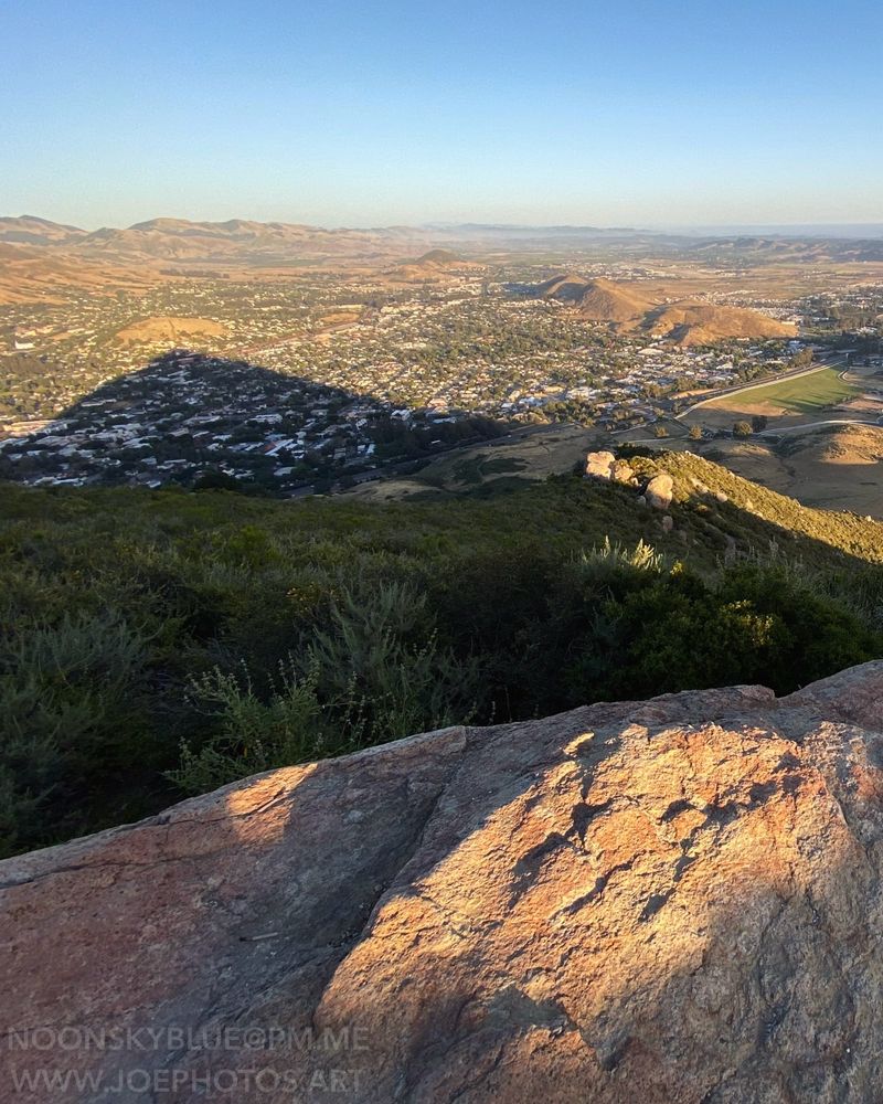 Sunlight illuminates the edge of a pink granite rock face in the foreground. A long shadow of the pointed hill on which the rock stands stretches over a small city below.