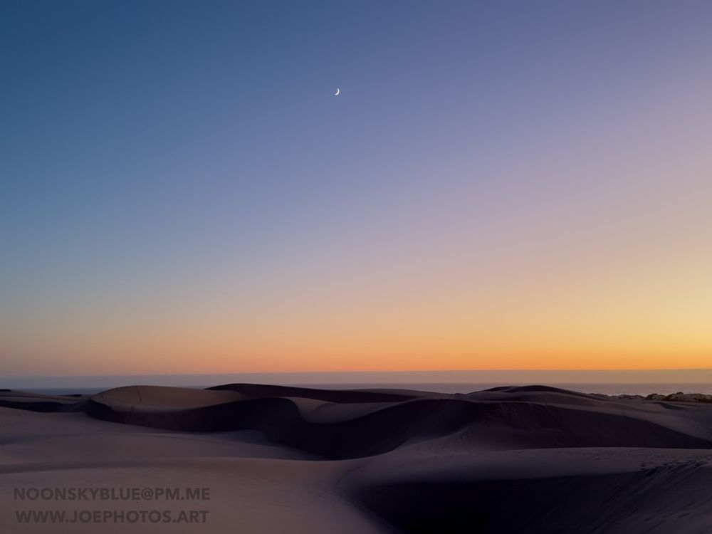 A crescent moon high above rolling sand dunes. Lingering light makes the sky orange, pink, and deep blue.