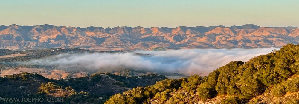 Low-lying evening fog drapes across a sunlit valley surrounded by rolling hills, with distant hills bathed in golden light under a clear blue sky.