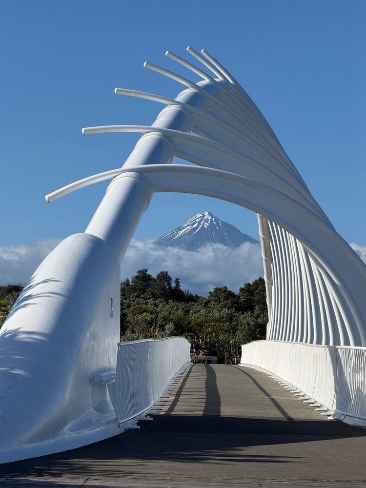Blue sky. A modern bridge with white rounded spikes similar to a rib cage. Through the opening of the spikes we see a triangular snow-capped mountain top. 