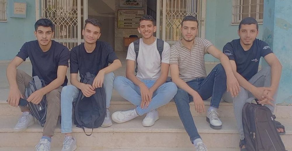 Mohammed with four friends from university. 
He’s wearing a striped polo shirt, jeans, and tennis shoes. They’re sitting on the stairs outside a building, smiling and relaxed.