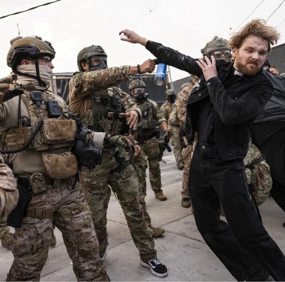 A young pastor wearing a white collar jumps out of the way as an Immigration and Customs Enforcement officer sprays pepper spray in his direction. The ICE officer is part of a group of several officers wearing what looks like military gear: camo uniform with masks, helmets, radios, guns and vests with pouches and pockets. The people are standing on what appears to be outdoor concrete flooring of some type.