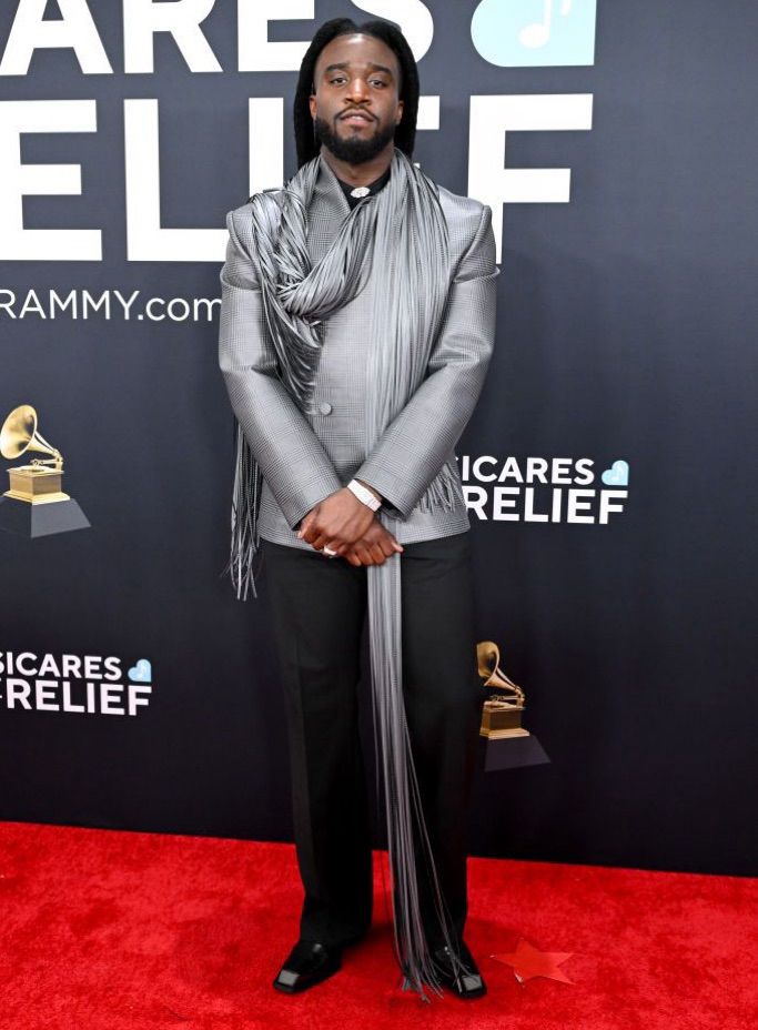 Shaboozey wears a shiny gray jacket with a long fringe scarf wrapped around his neck that hangs to the ground. With black pants and dress shoes.