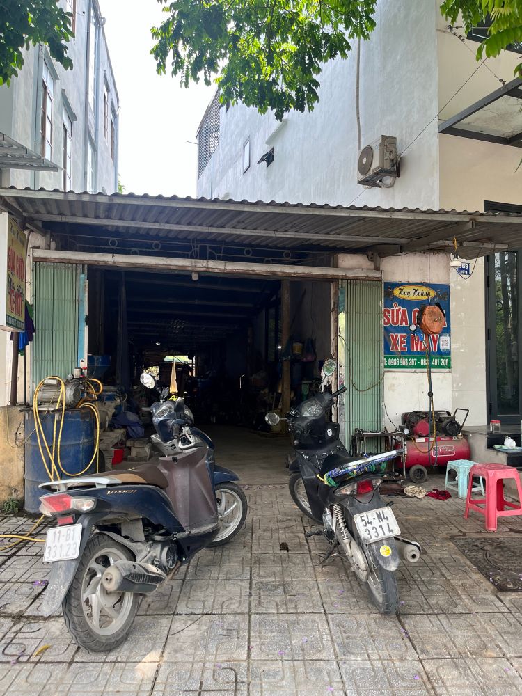 Two motorbikes parked on a tiled sidewalk outside a narrow, open garage filled with tools and equipment. A sign reading “Sửa chữa xe máy” (motorcycle repair) is mounted on the wall, indicating a Vietnamese motorbike repair shop.