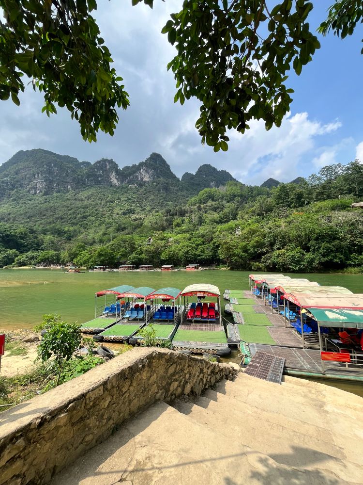 Covered wooden rafts with red and blue seats are docked along a riverbank under leafy tree branches, with misty mountains and dense forest in the background. A stone stairway leads down to the water’s edge.