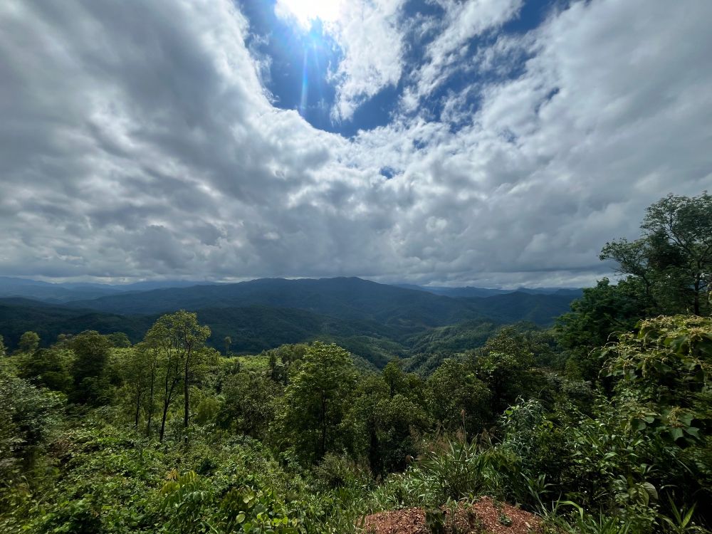 A dramatic landscape of forest-covered mountains stretches into the distance under thick, moody clouds breaking open to reveal a burst of sunlight.