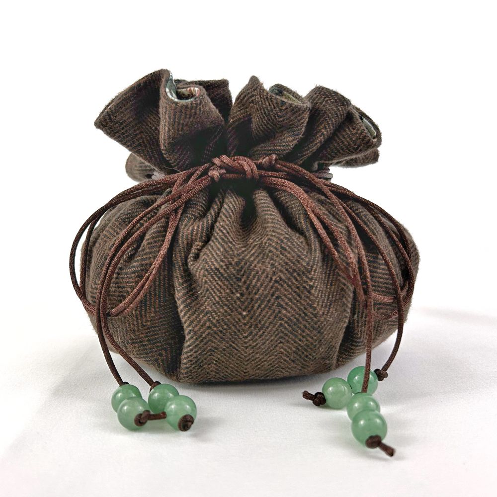 Exterior shot of a closed dice bag. The exterior of the bag is made of a brown and black woven herringbone flannel. The dice bag has brown synthetic cording used for the drawstrings. Each drawstring has two green stone beads tied on at the end. This is a picture of a medium size bag that holds approximately 140 dice. 